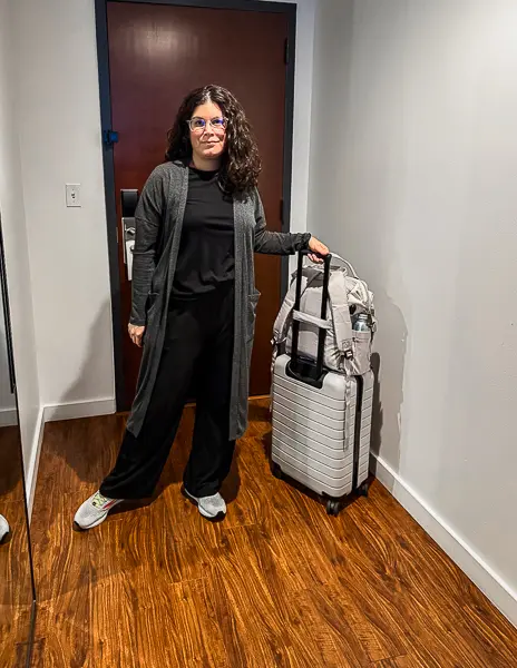Woman  (kathy) standing in a hotel hallway wearing a long gray cardigan, black outfit, and sneakers, holding a suitcase and backpack before embarking on an Alaska cruise