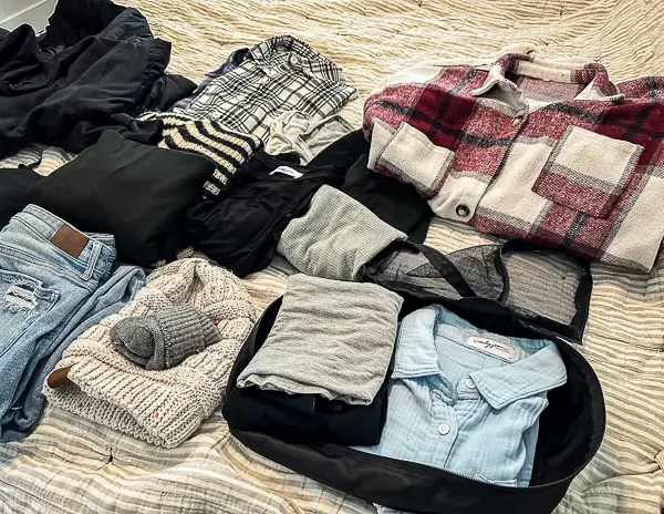 Flat lay of folded Alaska cruise clothing on a bed, including jeans, sweaters, fleece-lined leggings, a plaid shacket, knit hats, and a chambray shirt packed in a cube