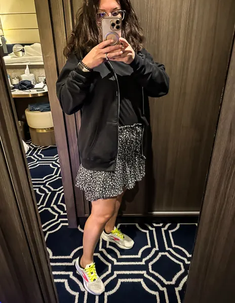 Woman wearing a black zip-up hoodie, black top, and floral ruffle short-skirt with sneakers, taking a mirror selfie in a cruise ship cabin before dinner on an Alaska cruise