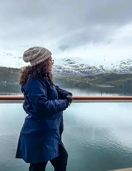 Woman (kathy) wearing a rain jacket, knit beanie, and gloves standing on a cruise ship balcony with snow-capped mountains and icy water in Glacier Bay, Alaska