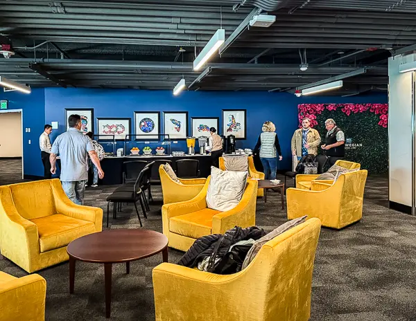 Haven priority embarkation lounge at the Seattle cruise port for Norwegian Cruise Line, featuring yellow chairs, a snack and beverage station, and a blue accent wall with framed artwork.