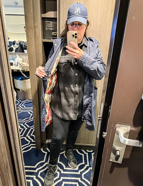 Woman (Kathy) wearing a black plaid flannel, rain jacket, black leggings, hiking sneakers, and a baseball cap in a cruise cabin mirror selfie before a rainy port day in Ketchikan, Alaska