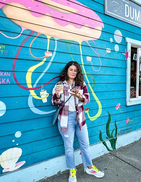 Woman (kathy) wearing a red plaid shacket, jeans, and sneakers while holding coffee and a donut in front of a colorful jellyfish mural in Ketchikan, Alaska