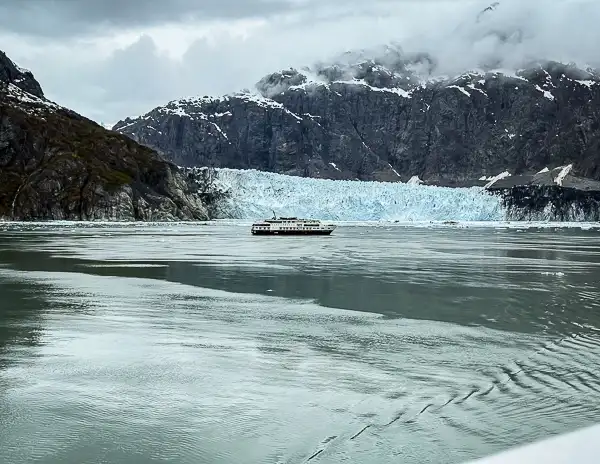 National Geographic expedition ship sailing near Margerie Glacier in Glacier Bay, Alaska, with icy blue glacier face and rugged mountain backdrop.