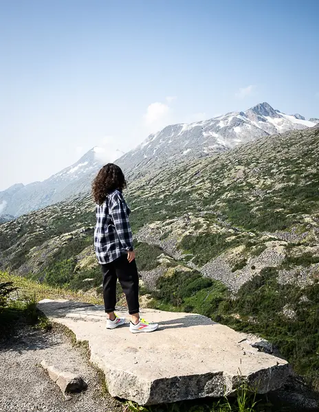 Woman (kathy) wearing a navy plaid button-down and black pants standing on a rock ledge overlooking a scenic mountain view near Skagway, Alaska