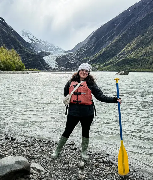 Woman (kathy) in a life vest and knit cap holding a paddle while standing in front of a glacier lake near Skagway, Alaska, wearing rain boots and layered cold-weather gear.