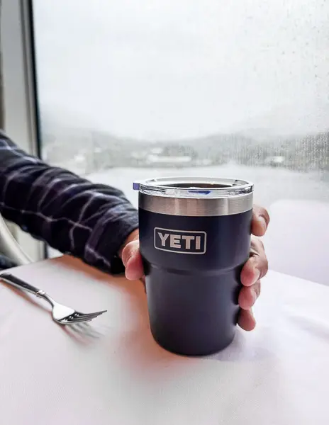 Hand holding a YETI coffee mug by the window on a cruise ship, with a rainy view outside.