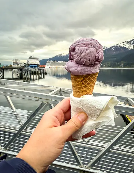 Hand holding a waffle cone with huckleberry ice cream in front of a cruise port in Juneau, Alaska, with mountains and ships in the background.