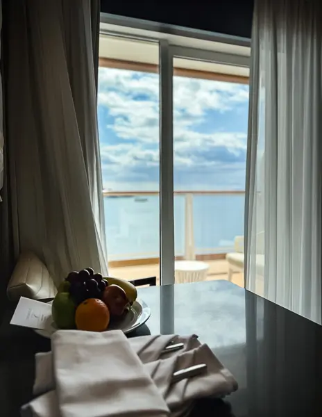 View through sheer curtains looking out from The Haven Penthouse Suite 17112 on Norwegian Encore, showing the private balcony and ocean beyond with fruit and napkins in the foreground.