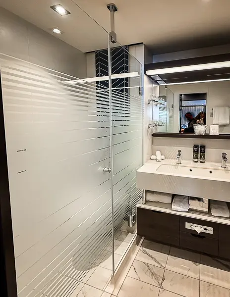 Bathroom inside The Haven Penthouse Suite 17112 on Norwegian Encore, featuring a frosted glass walk-in shower, dual sink vanity, shelves with towels, and a large mirror.