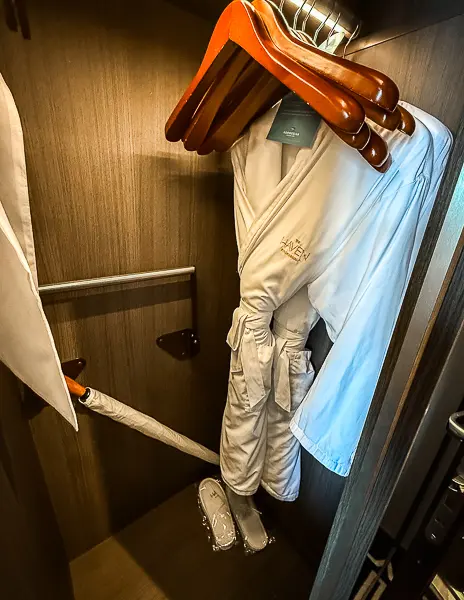 Open closet in The Haven Penthouse Suite 17112 on Norwegian Encore, showing a hanging robe embroidered with The Haven logo, wooden hangers, an umbrella, and a pair of slippers.