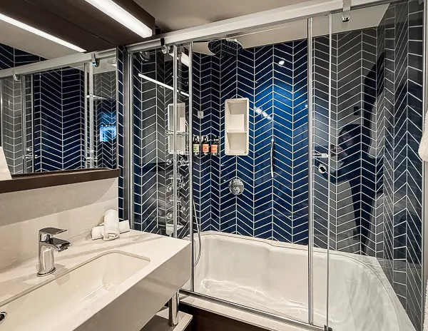 Bathroom of NCL Encore Haven Penthouse Suite 17112 featuring a tub, rainfall shower, and blue chevron tile.