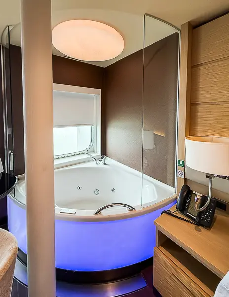 Corner jacuzzi tub with glowing blue lights in The Haven Suite 14138 on Norwegian Getaway, next to a window and bedside table.
