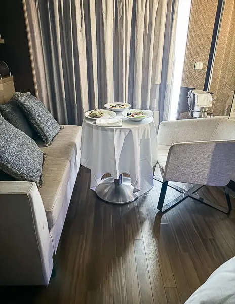 In-room dining setup in Norwegian Getaway’s Haven Suite 14138, with a white tablecloth, plates of food, and a sofa and chair near the balcony.
