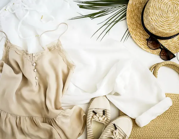 Flat lay of a light cruise outfit with a tan dress, straw hat, espadrilles, sunglasses, and beach bag.