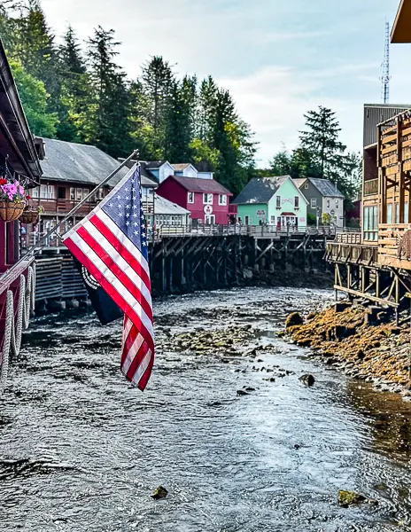 American flag hanging along Creek Street in Ketchikan, Alaska, with colorful historic buildings on stilts lining the creek at low tide.