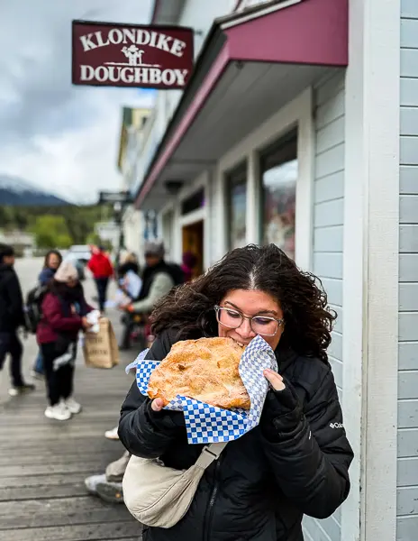 Woman (Kathy) holding and eating a large piece of fry bread in front of Klondike Doughboy in Skagway, Alaska, with other people and shops in the background.