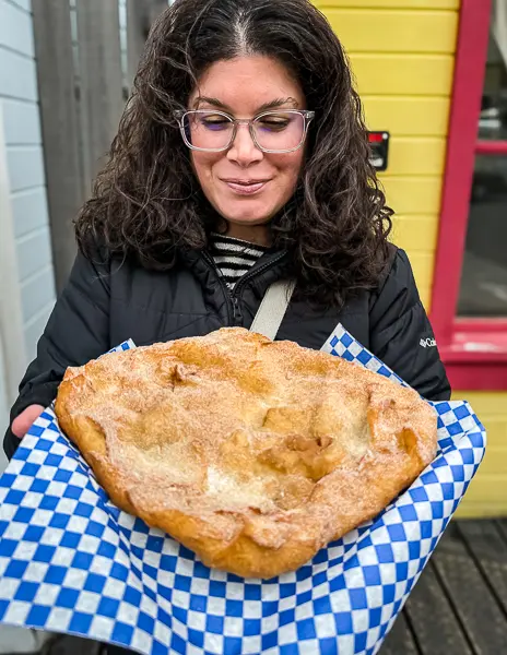 Woman (Kathy) smiling while holding a large piece of cinnamon sugar fry bread on blue checkered paper outside Klondike Doughboy in Skagway, Alaska.