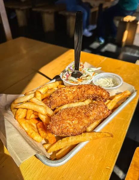 Tray of Alaskan halibut fish and chips served with fries, coleslaw, and tartar sauce on a wooden table in a casual restaurant.