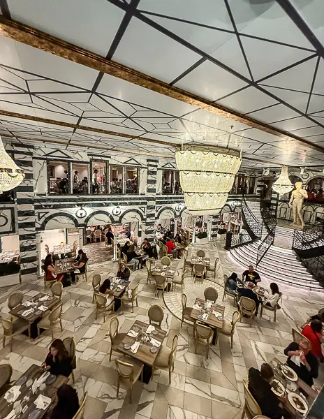 View of the elegant main dining room on Carnival Firenze, with chandeliers, marble-patterned floors, and guests dining at tables.