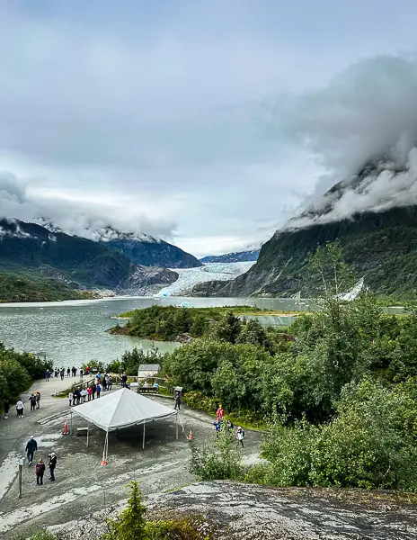 Visitors gathered at a scenic viewpoint overlooking Mendenhall Glacier in Juneau, Alaska, surrounded by mountains and misty clouds.