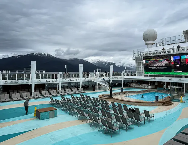 Norwegian Encore pool deck on a cool May sea day to Alaska, with lounge chairs mostly empty under cloudy skies and snow-capped mountains in the background.