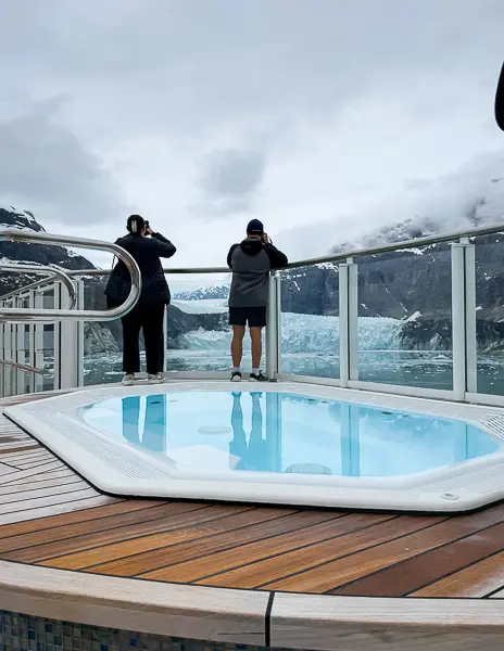 Guests standing by the railing with binoculars near a hot tub on the Haven sundeck of Norwegian Encore, overlooking Glacier Bay in Alaska