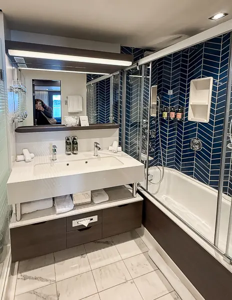 Bathroom inside the Haven Penthouse Suite on the Norwegian Encore, showing the sink area, L'Occitane toiletries, and full tub with glass shower doors.