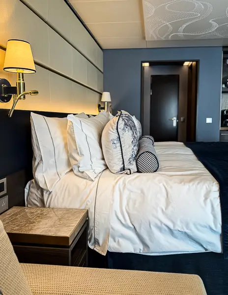 King bed inside our Haven Penthouse suite on the Norwegian Encore, with crisp white linens, plush pillows, and modern lighting.