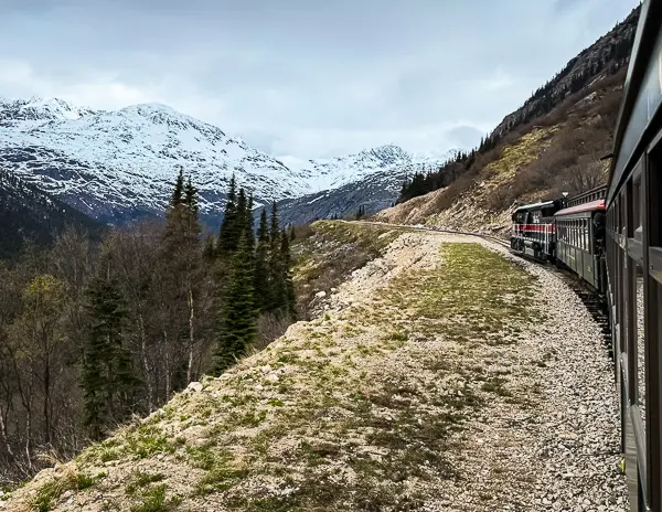 Scenic view from the White Pass and Yukon Route train in Skagway, Alaska, with snow-covered mountains and forested slopes in the background.