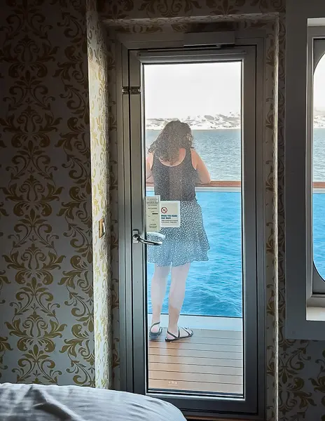 View from inside Carnival Firenze Ocean Suite 7359 looking out through the balcony door, with a guest standing outside facing the ocean.