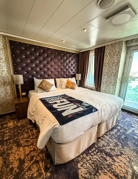 King-size bed in Carnival Firenze Ocean Suite 7359 with patterned carpet, ornate wallpaper, and balcony door leading outside.