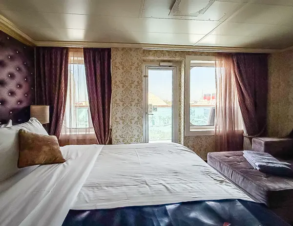 Carnival Firenze Ocean Suite 7359 with king-size bed, purple curtains, and balcony door with two picture windows overlooking the water.