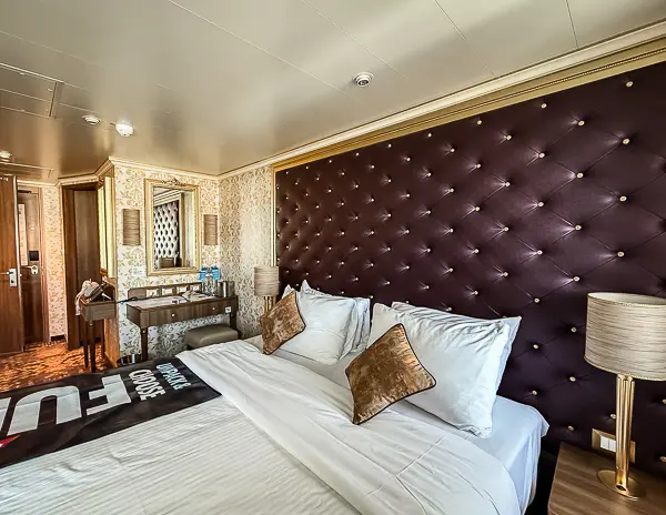 Carnival Firenze Ocean Suite 7359 showing the king bed with tufted headboard and the vanity desk with mirror in the background.