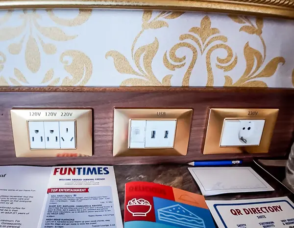 A close-up of the desk outlets in Carnival Firenze Ocean Suite 7359, showing two U.S. (120V) outlets, one 220V outlet, one European outlet, and two USB-A ports.
