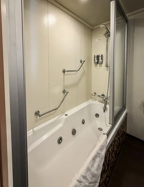 Large jacuzzi tub with shower combo in Carnival Firenze Ocean Suite 7359, featuring grab bars, glass shower doors, and mounted dispensers.