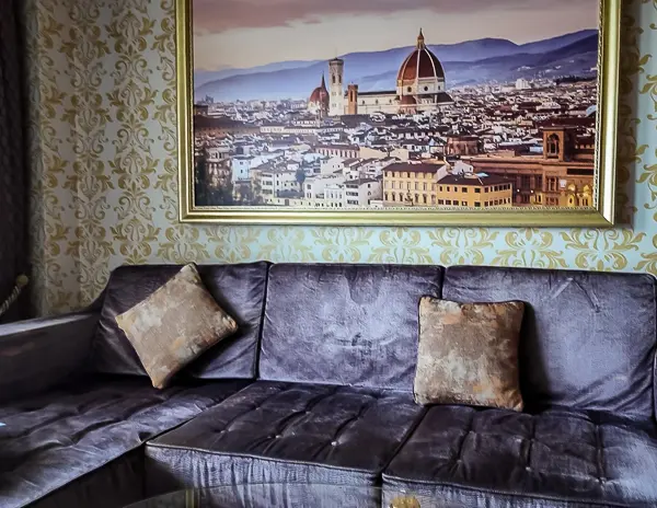 Large gray sofa in Carnival Firenze Ocean Suite 7359 with gold-accent pillows, set against ornate wallpaper and a framed painting of Florence.
