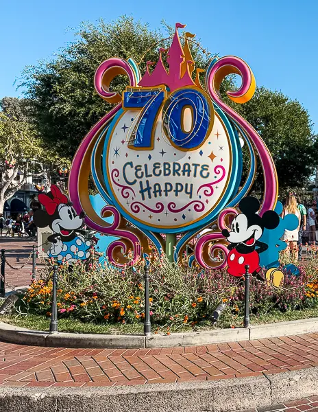 Disneyland 70th anniversary sign with “Celebrate Happy” message, featuring Mickey and Minnie Mouse surrounded by colorful flowers in Town Square.