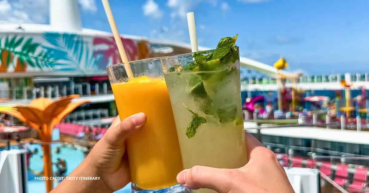 Two hands holding drinks, one mango smoothie and one mojito with mint, raised in a toast by the pool deck of a Royal Caribbean cruise ship on a sunny day.