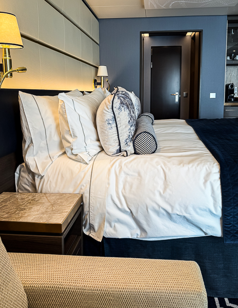 Cruise ship cabin nightstand next to the bed with lamp and pillows, ideal for storing small items like chargers, lip balm, or a Kindle.