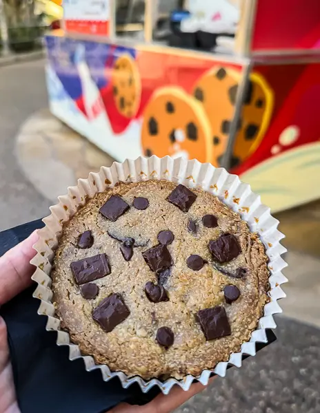 Hand holding a warm Jack-Jack Cookie Num Num with chocolate chunks in front of the cookie cart at Disney California Adventure.
