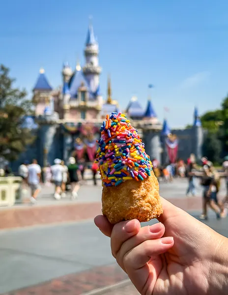 Hand holding a Matterhorn Macaroon topped with blue icing and rainbow sprinkles, with Sleeping Beauty Castle in the background at Disneyland.