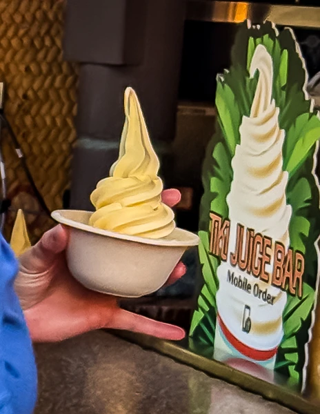 A hand holding a bowl of pineapple Dole Whip soft serve in front of the Tiki Juice Bar sign at Disneyland.