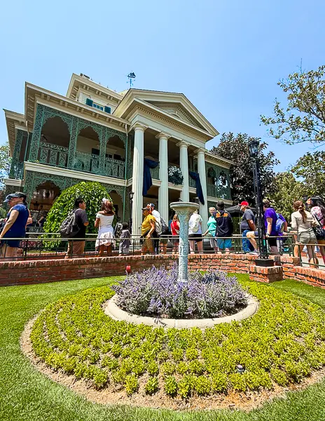 Guests waiting in line outside the Haunted Mansion at Disneyland on a sunny day.