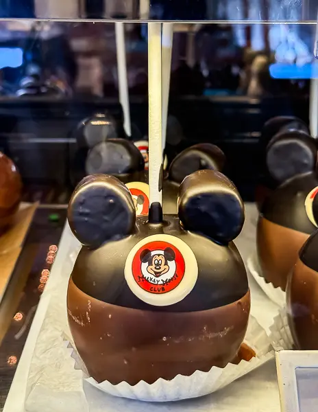 Mickey Mouse chocolate-dipped apple decorated with ears and a Mickey Mouse Club logo, displayed in a glass case at a Disneyland candy shop.