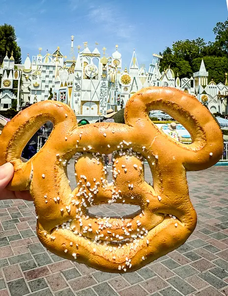 Hand holding a Mickey-shaped pretzel in front of the "it’s a small world" attraction at Disneyland