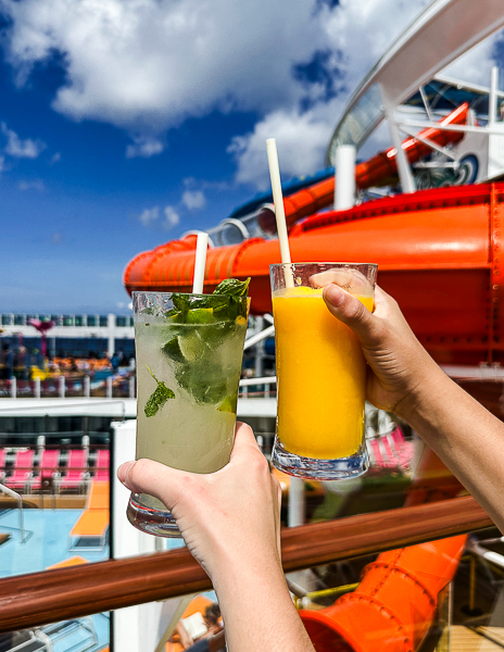 Two cocktails held up on the pool deck of a Royal Caribbean cruise ship, one mojito and one frozen mango drink, with waterslides in the background