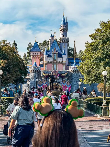 Guest wearing Mickey-shaped pretzel ears walking toward Sleeping Beauty Castle at Disneyland.