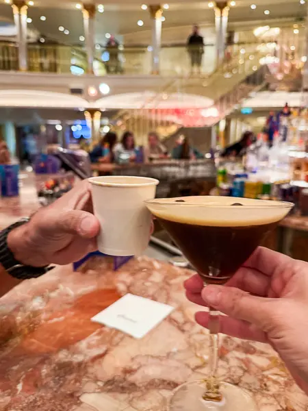 Two guests clinking drinks at a Carnival cruise ship bar, one holding an espresso martini and the other a double shot of espresso, both covered by Carnival drink packages.