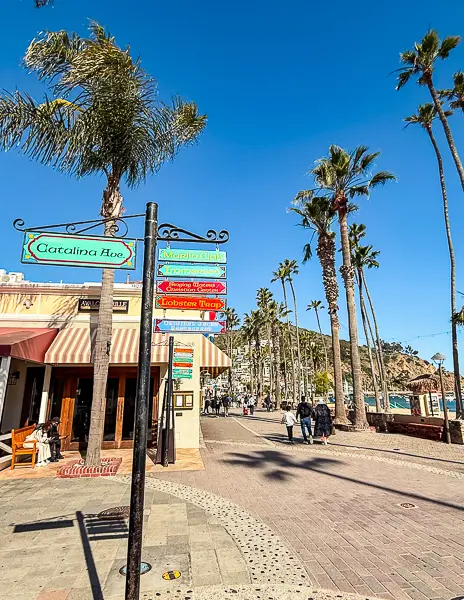 Street view of Catalina Avenue in Avalon, Catalina Island, with a colorful signpost, palm trees, and shops along the waterfront.